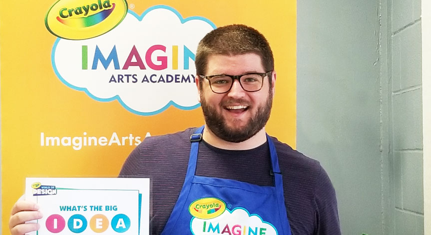 A Crayola Imagine Arts Academy art instructor holding a certificate standing in front of a Crayola Imagine Arts Academy banne