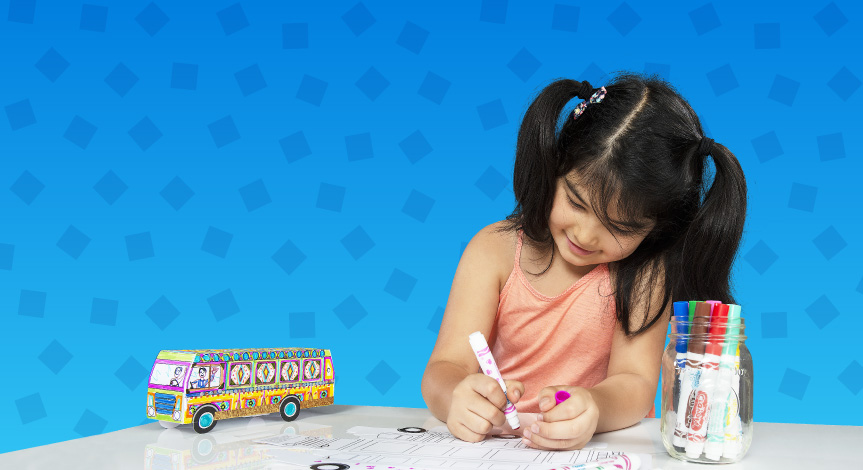 Girl coloring a Crayola Imagine Arts Academy bus take home while a completed version is next to her on the table. Image is on